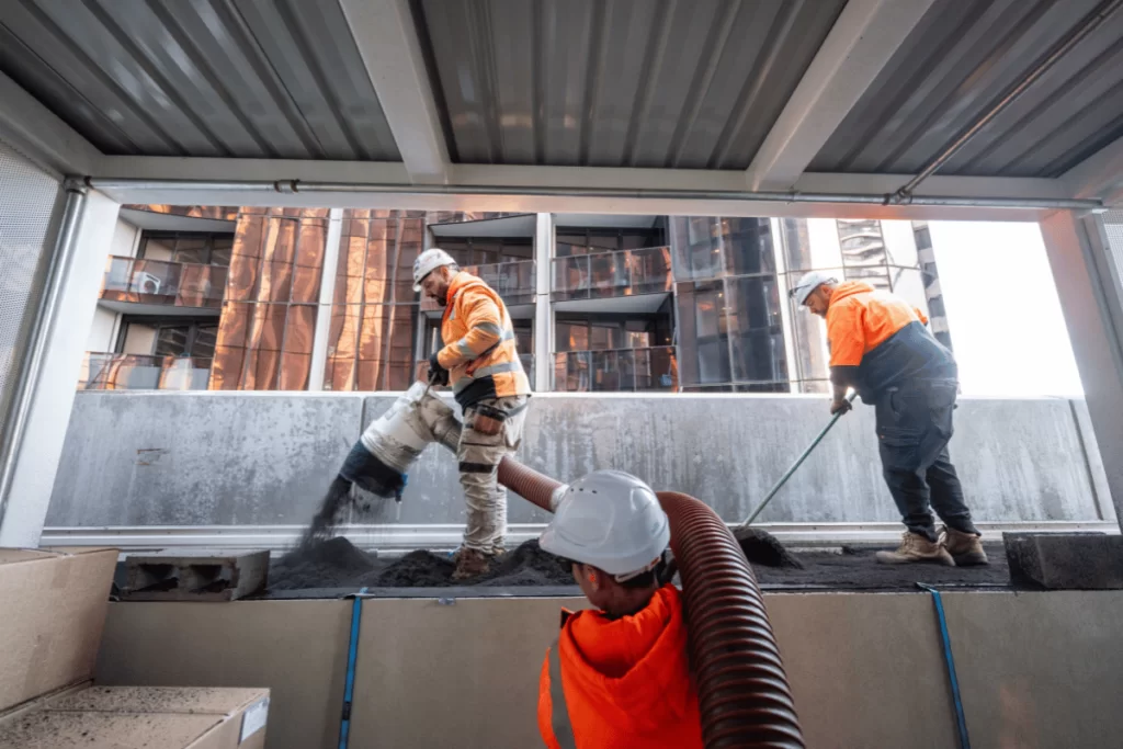 On the 9th story of a city building, three workers in orange high-vis are using a blower to install soil