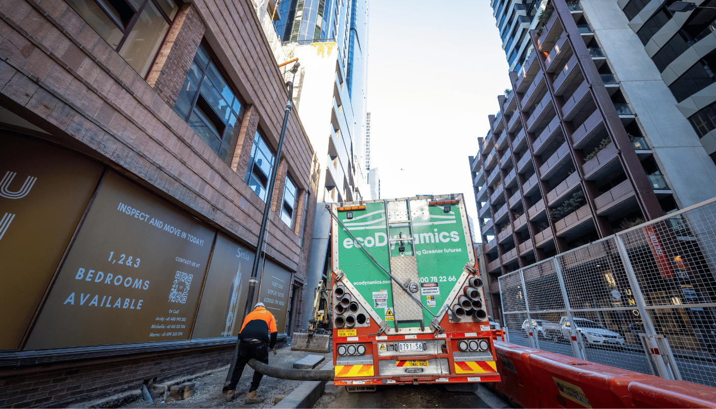 An ecoDynamics blower truck is parked on a city street between high rise buildings. While a worker is high-vis is pulling out a blower hose from the truck.