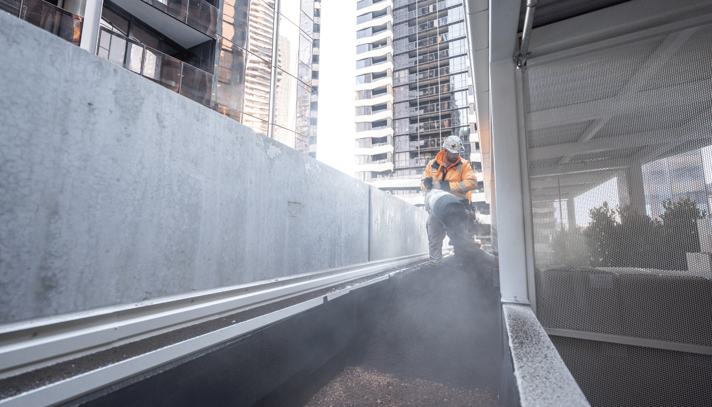 An ecoDynamics worker is holding a blower hose from a blower truck and blowing soil on top the ground of a 9 level apartment.