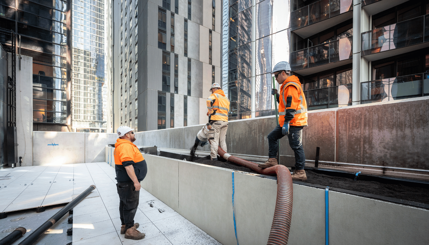 Three ecoDynamics landscaping workers in high-vis are using a blower hose to blow soil. One is holding a broom while talking to the supervisor.