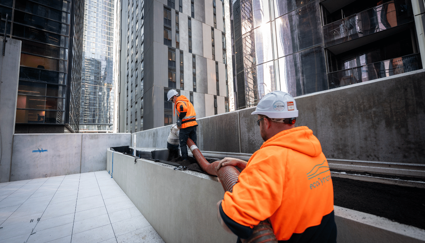 On the 9th level of a building in the city two ecoDynamics workers are using a blower hose to blow soil into a long planter box.