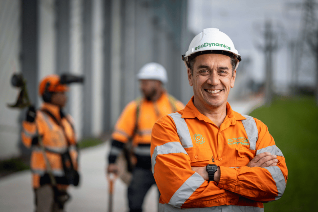 An ecoDynamics male worker standing with crossed arms wearing high vis and a white hard hat smiling at the camera.